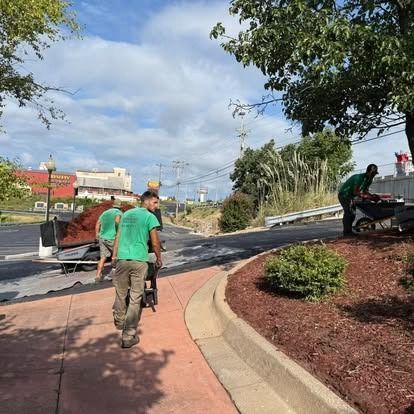Landscapers in green shirts spreading mulch near a sidewalk under a cloudy sky.