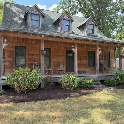 Wooden cabin with a porch and three dormer windows. Surrounded by shrubs, a lawn, and trees.