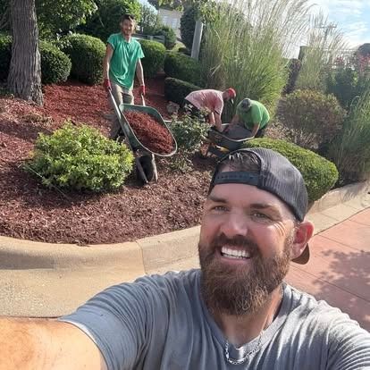 Man taking selfie with landscaping crew mulching flowerbeds; sunny day.