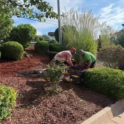 Two men spreading mulch in a landscaped area with shrubs, tall grass, and a building, outdoors.