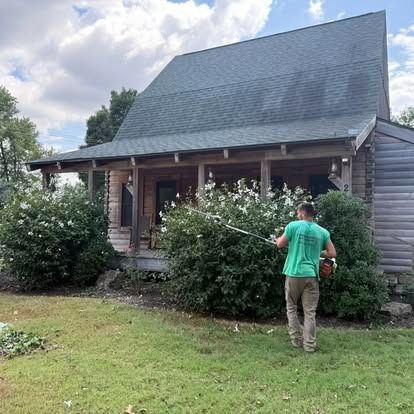 Man trimming a flowering bush in front of a cabin.