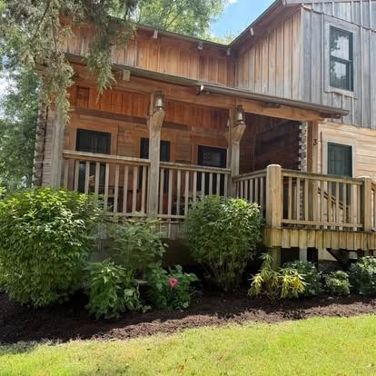 Wooden cabin with porch and landscaping; green bushes and grass in the foreground.