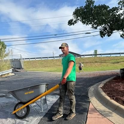 Man in green shirt pushing a wheelbarrow on asphalt near landscaping.