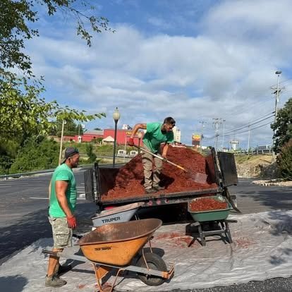 Two men spreading red mulch from a truck into a wheelbarrow; sunny day.