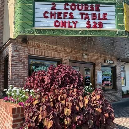 Restaurant exterior with a sign for a $29, 2-course chef's table. Red brick, green accents, and colorful plants.