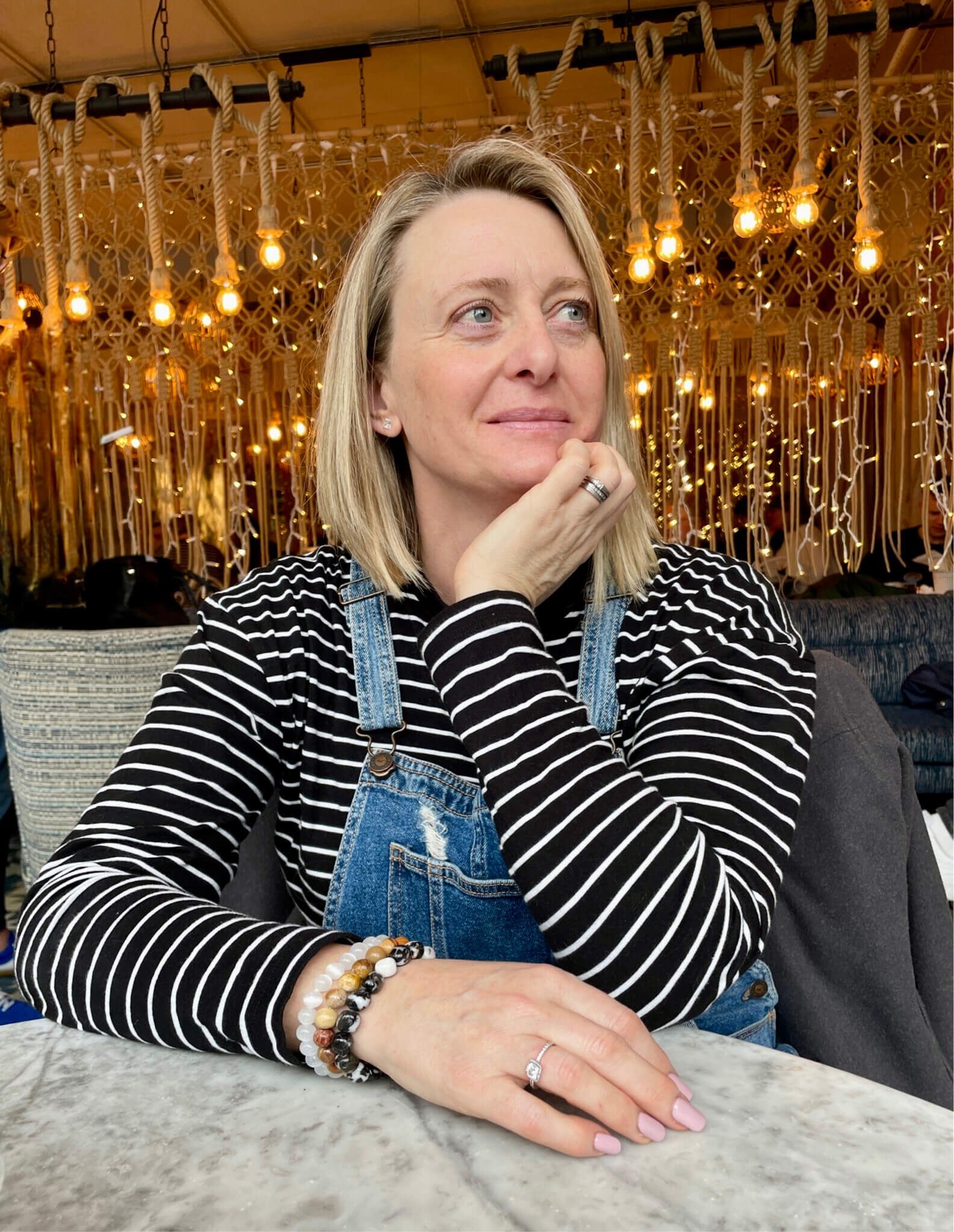 Michelle Winzar  is sitting at a table wearing overalls and a striped shirt.
