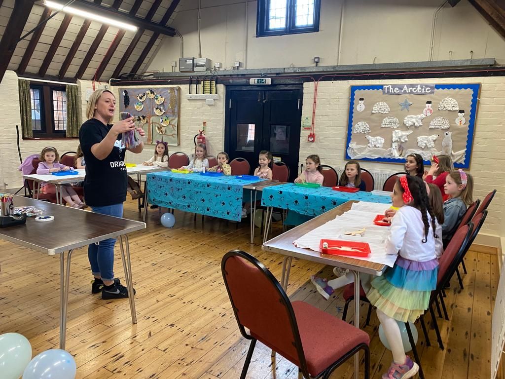 A woman is standing in front of a group of children sitting at tables in a room at Dragonfly-craft parties.
