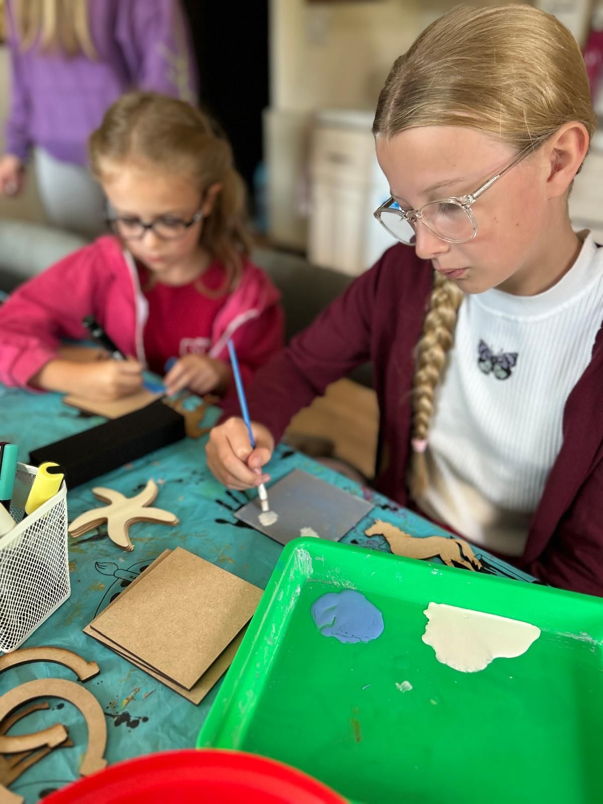 Two young girls are sitting at a table painting on cardboard  at Dragonfly crafts parties.