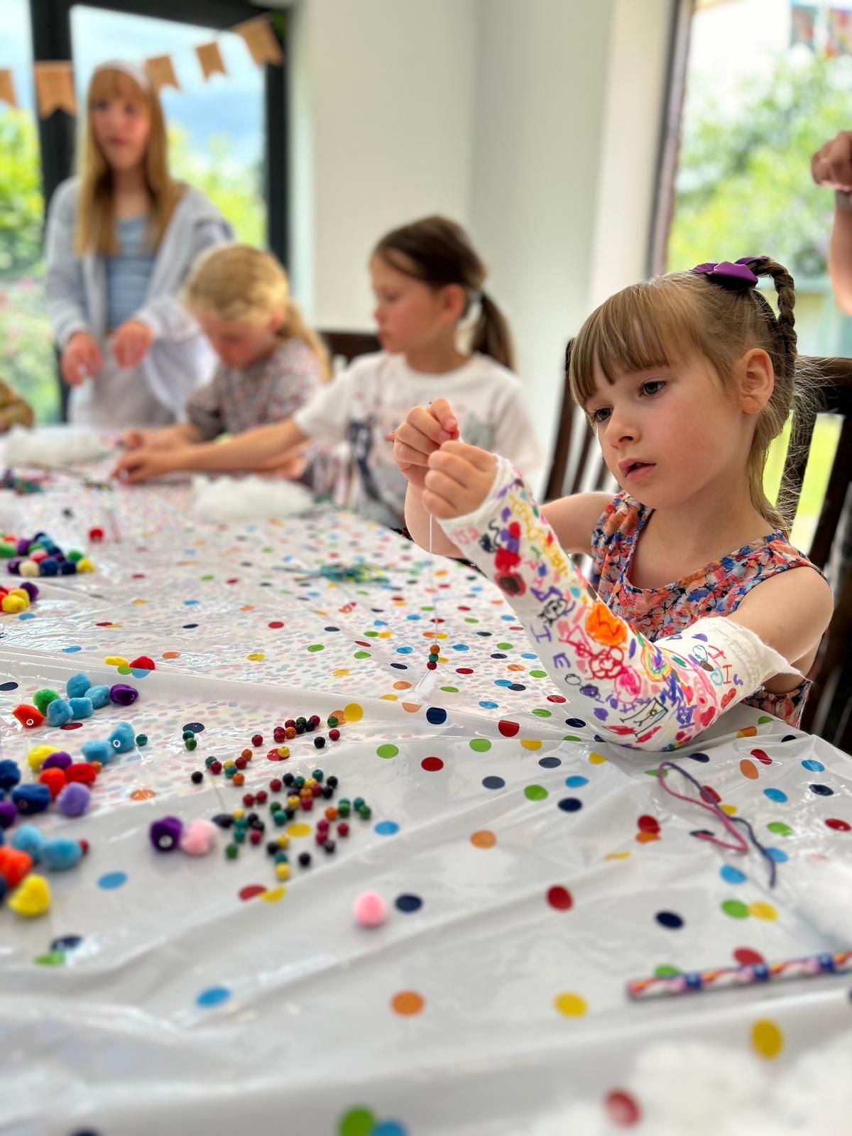 A little girl with a cast on her arm is sitting at a table with beads at Dragonfly-craft parties.
