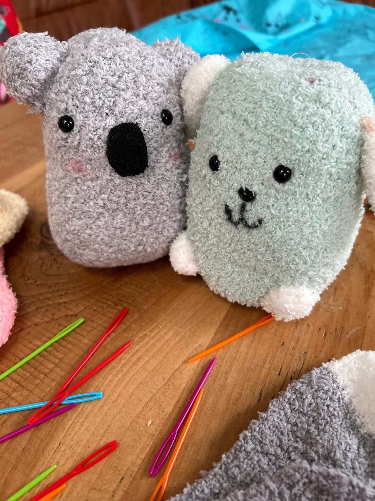 Two stuffed koalas are sitting on a wooden table next to knitting needles, for  Dragonfly-craft parties