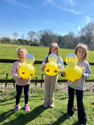 Three children holding yellow paper lanterns in a sunny grassy field