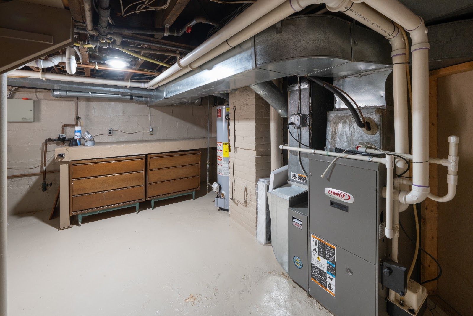 Basement utility room with furnace, ductwork, and storage cabinets under exposed pipes.