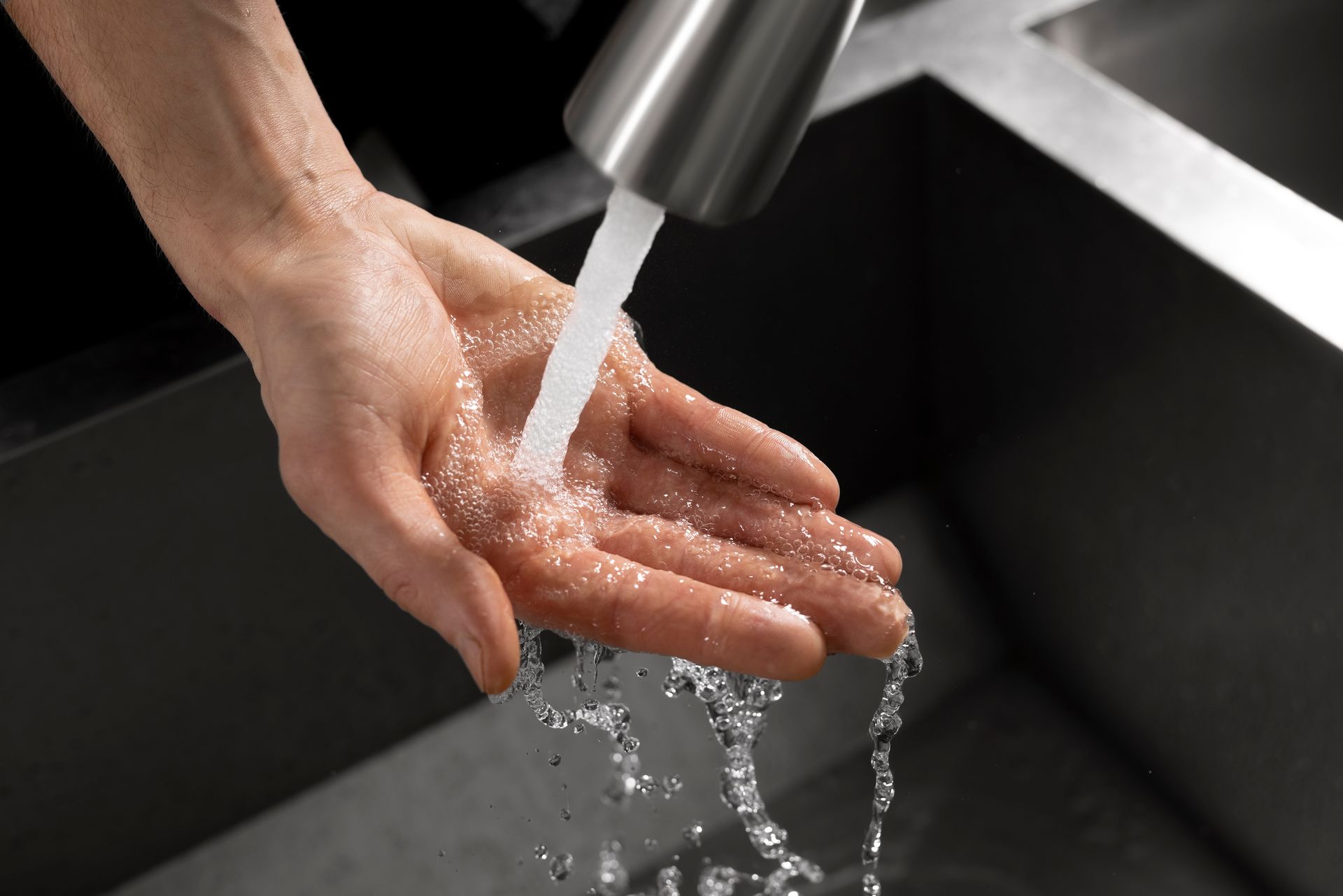 Hand under running faucet, washing with water splashing into a dark sink.