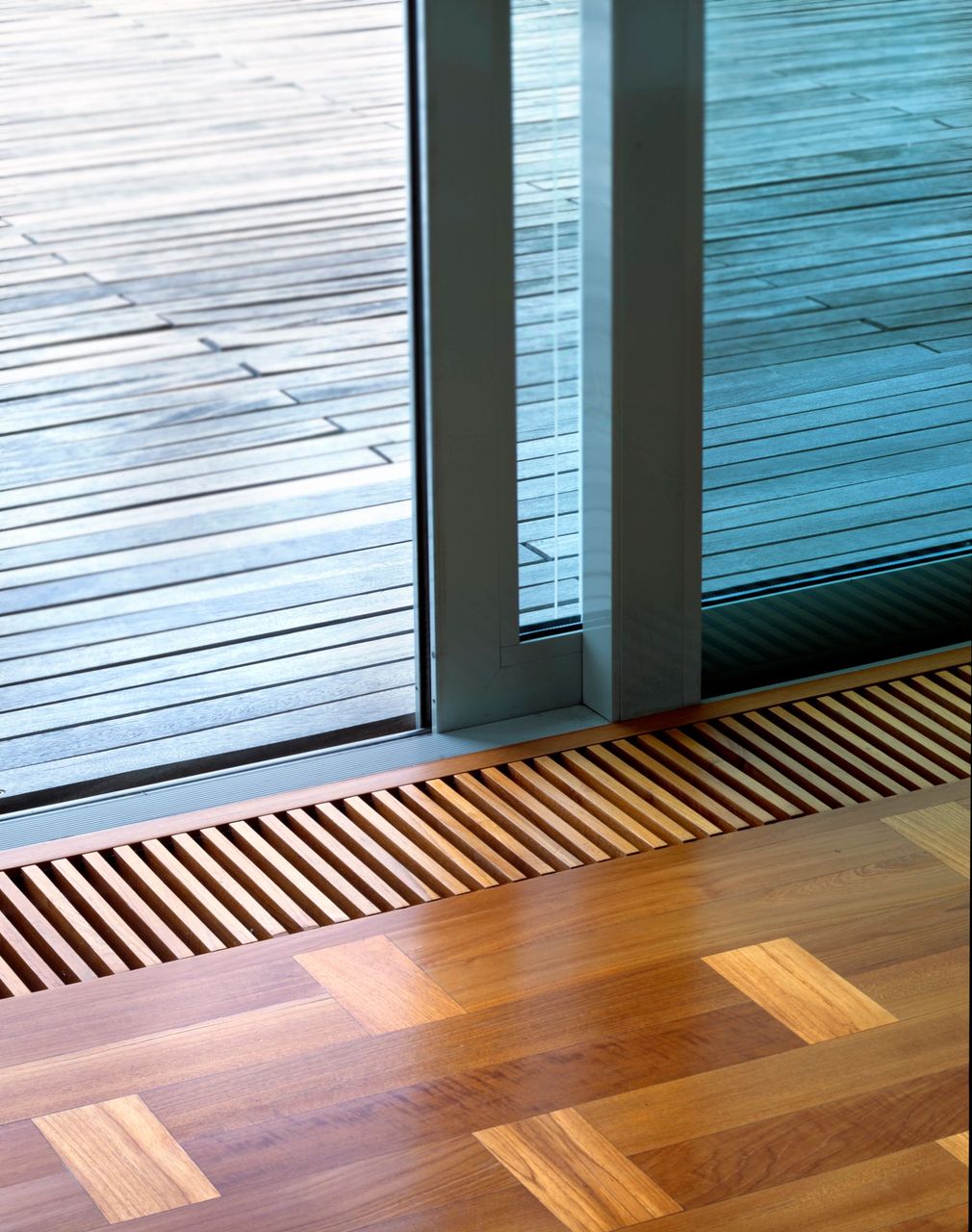 Pavimento in legno accanto a porte a vetri con bocchetta di ventilazione in metallo e pavimentazione esterna a strisce oltre.