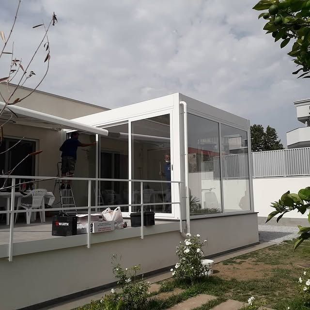 Patio bianco chiuso annesso a una casa, con ringhiera e cielo nuvoloso, accanto a un cortile e arbusti da giardino.