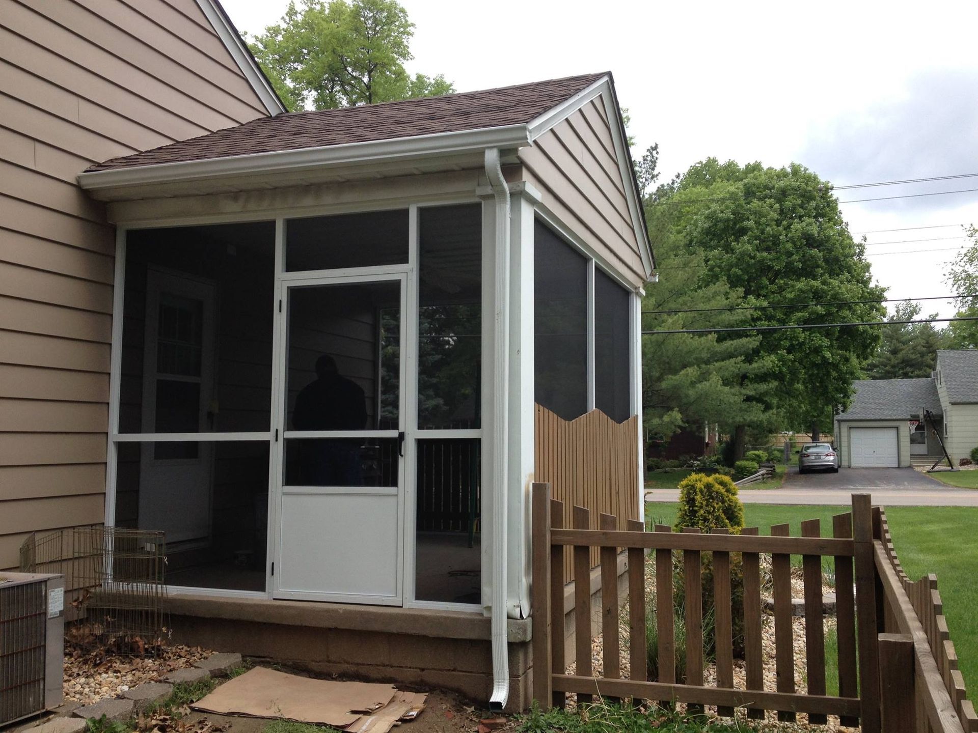 Porch with white screen door and brown railing beside a beige house, with a yard and street beyond.