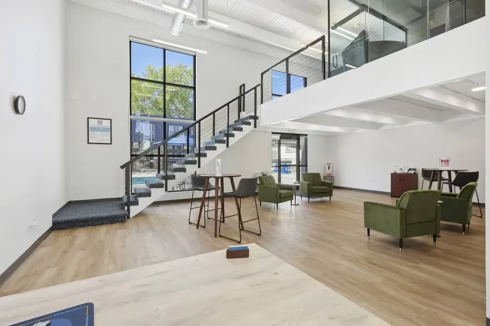 A bright, modern lobby with light wood floors, a central staircase, tall windows, and several olive-green armchairs.
