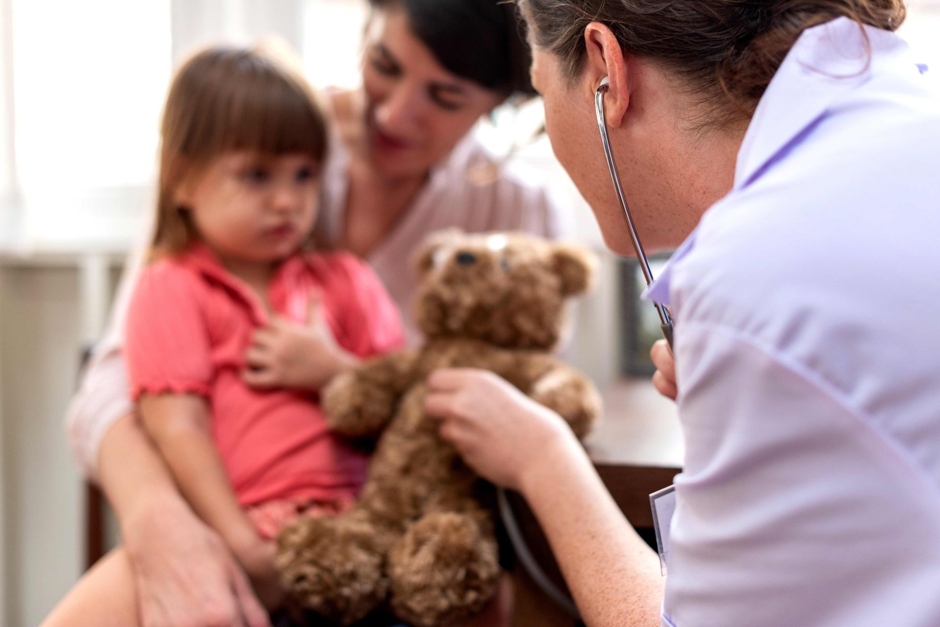 Doctor examining a child holding a teddy bear, with a parent nearby Doctor examining a child holding a teddy bear, with a parent nearby