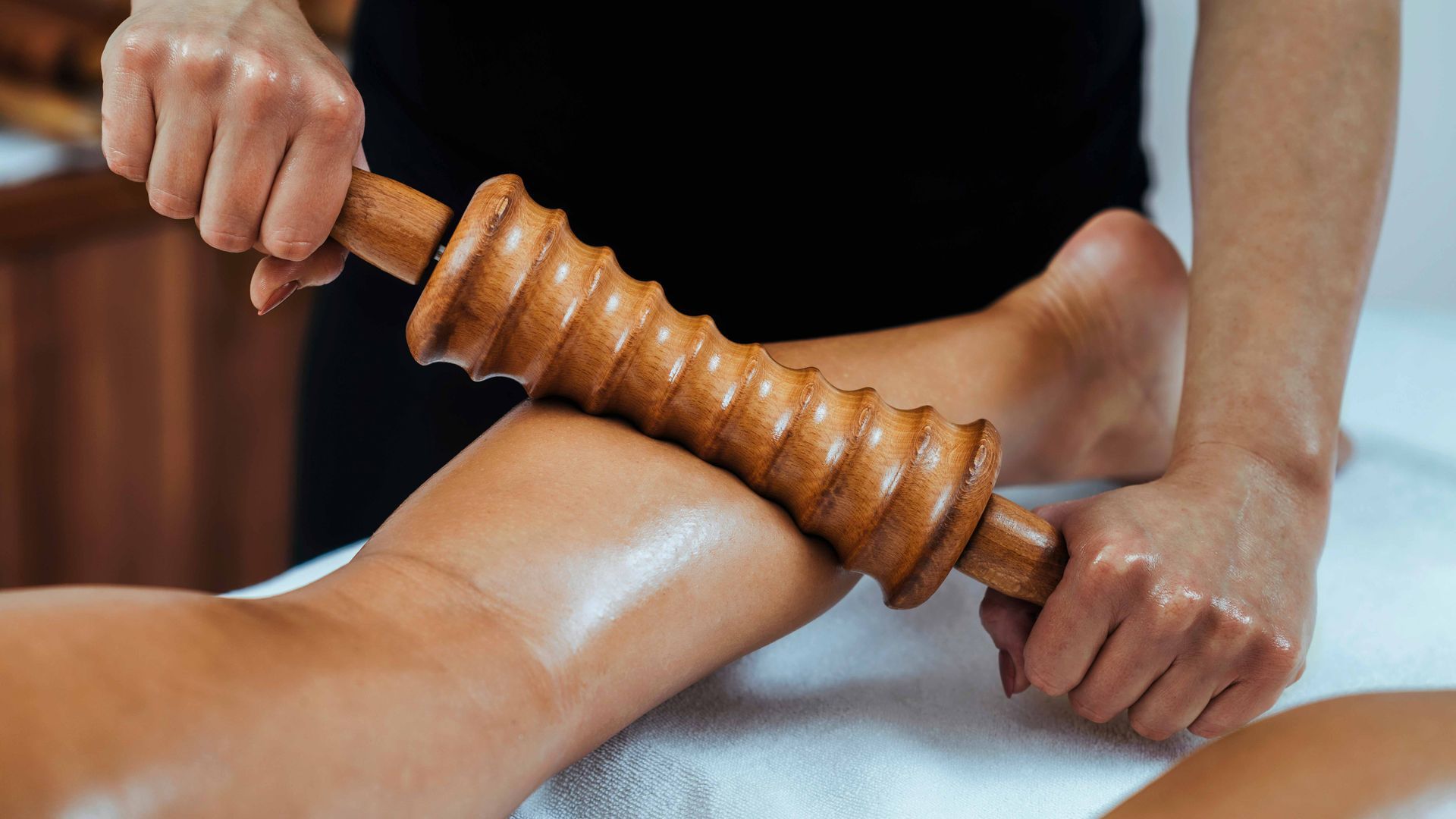 Hands using a wooden massage roller on a person’s calf over a white towel