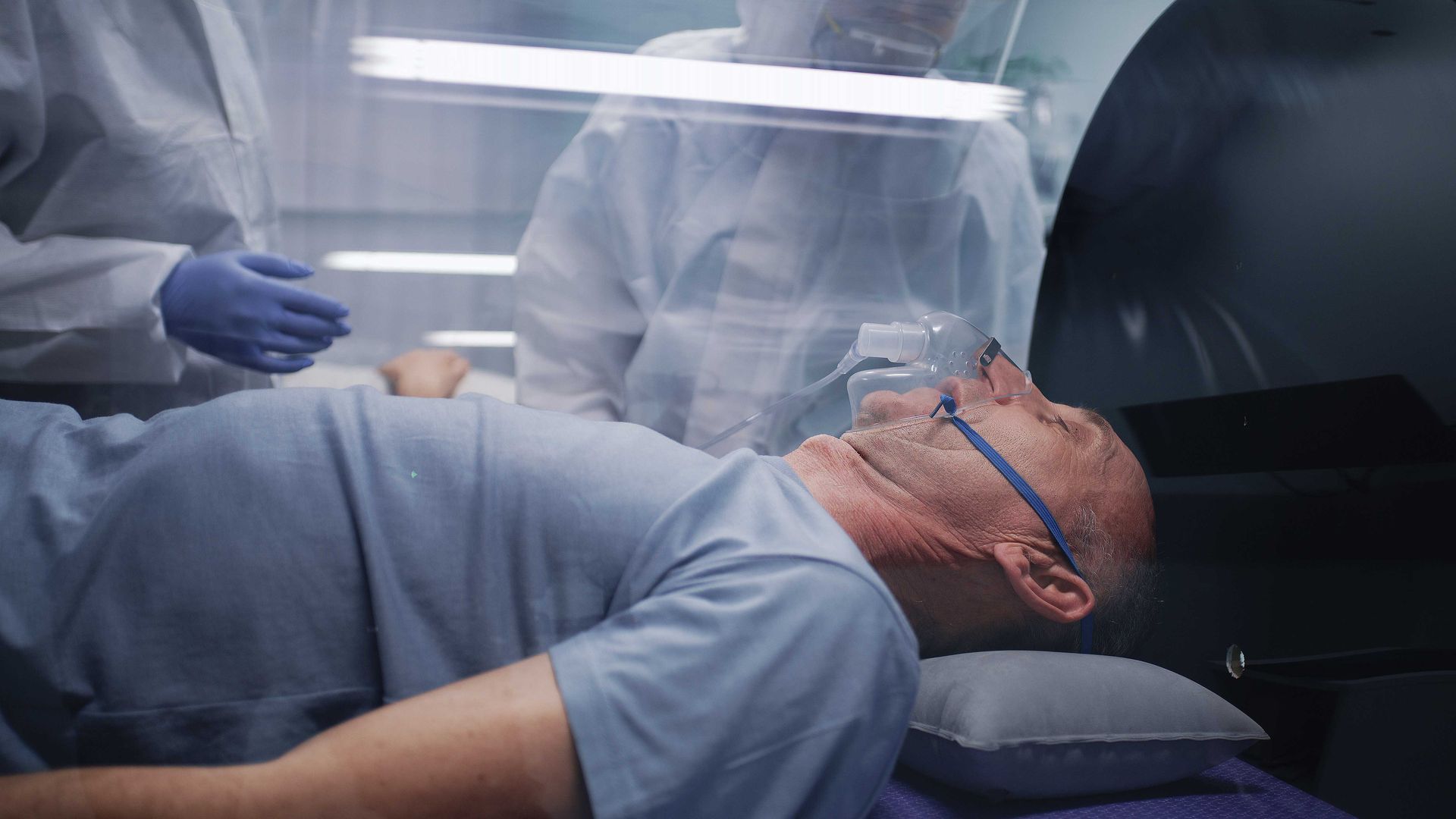 Patient lying on a hospital bed under a clear isolation tent, wearing an oxygen mask. Patient lying on a hospital bed under a clear isolation tent, wearing an oxygen mask.