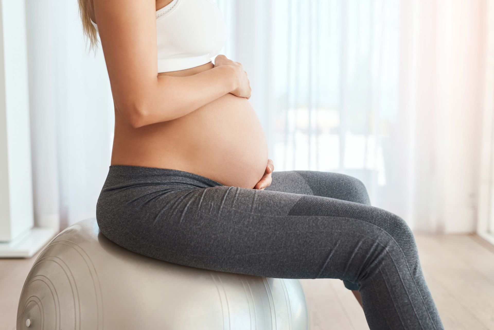 Pregnant person sitting on a white ottoman in a bright room, side profile with belly visible