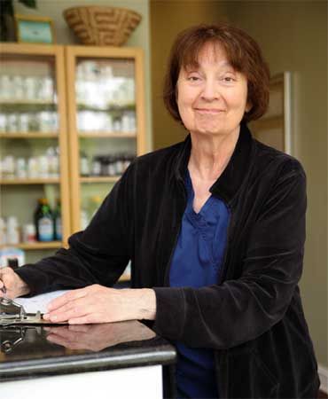 Person standing in a kitchen, leaning on a counter with shelves of dishes behind them.
