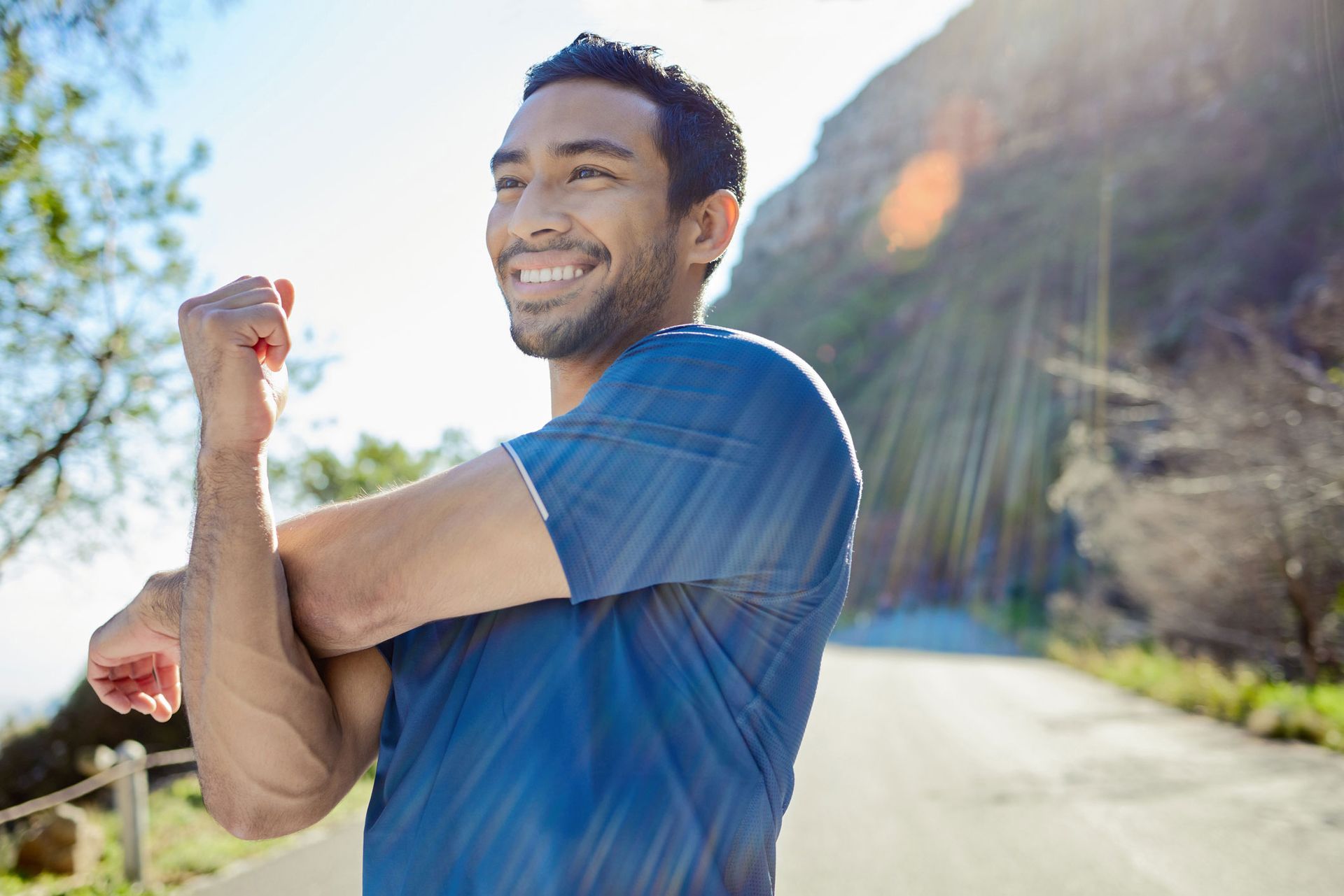 Smiling man stretching his arm outdoors on a sunny road with mountains in the background