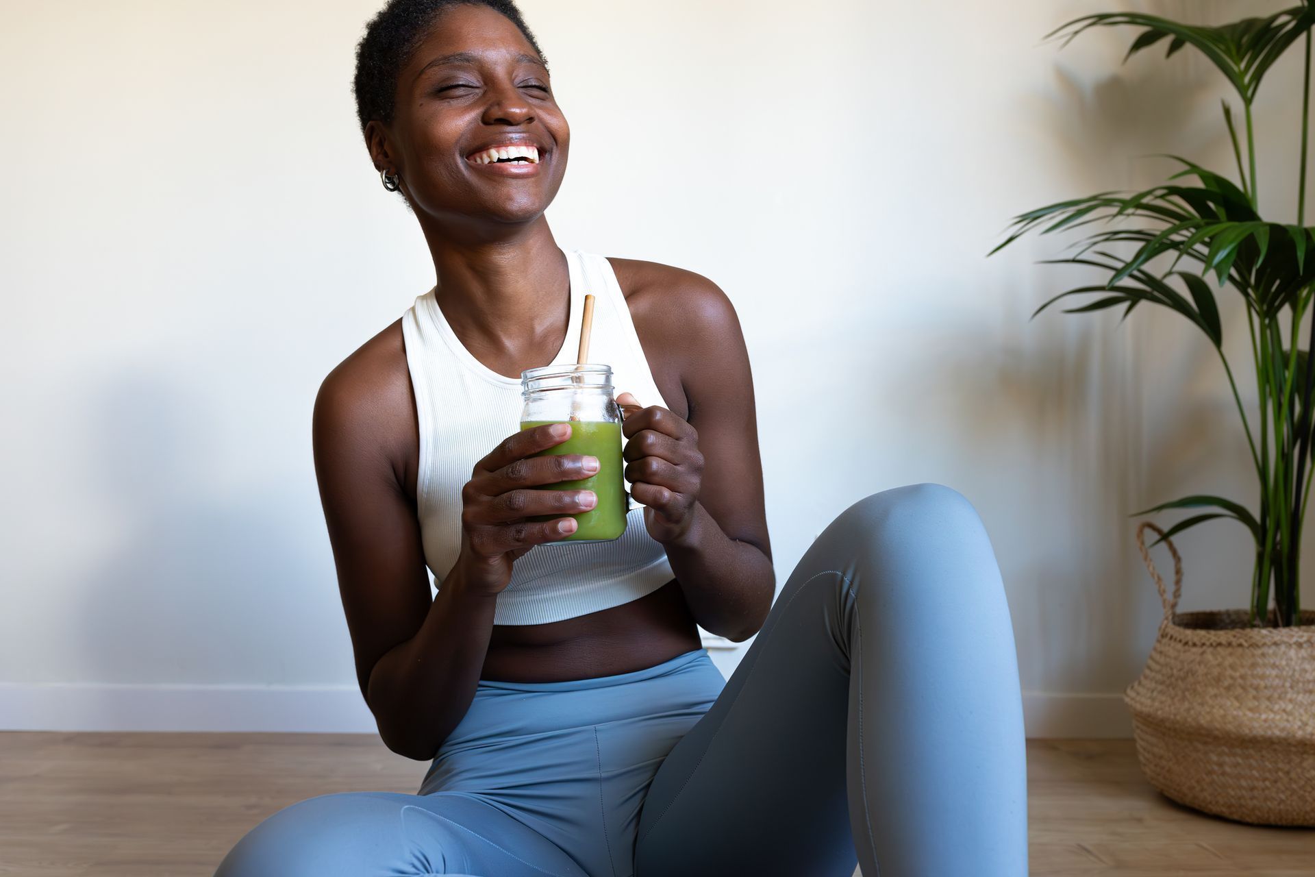 Smiling person in workout clothes holding a green smoothie on a yoga mat beside a potted plant Smiling person in workout clothes holding a green smoothie on a yoga mat beside a potted plant