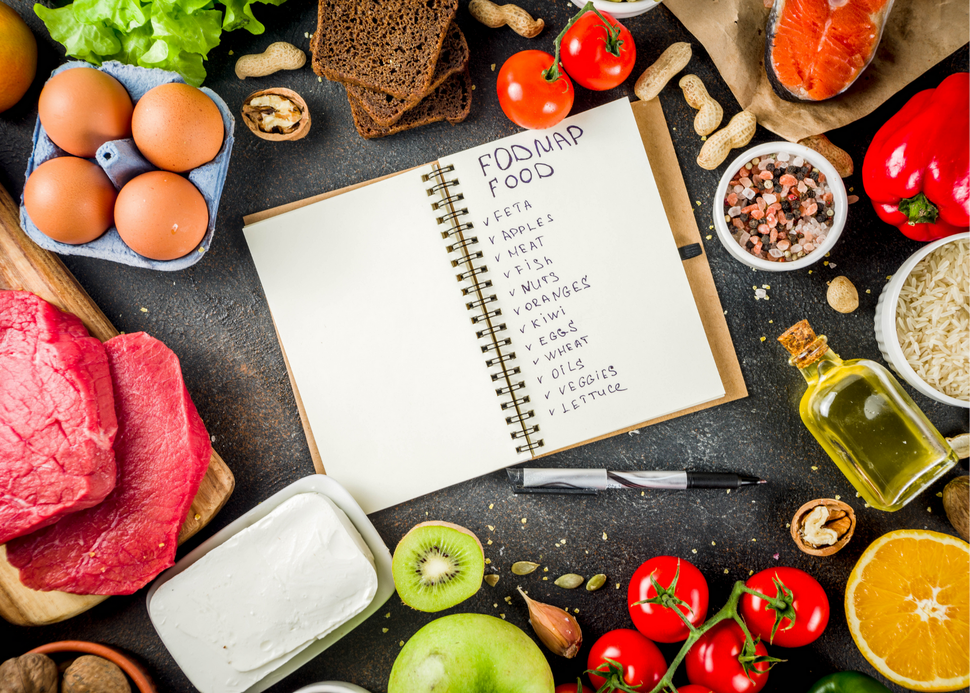 Open cookbook surrounded by fresh ingredients, eggs, bread, kiwi, tomatoes, meat, oil, and spices on a dark table