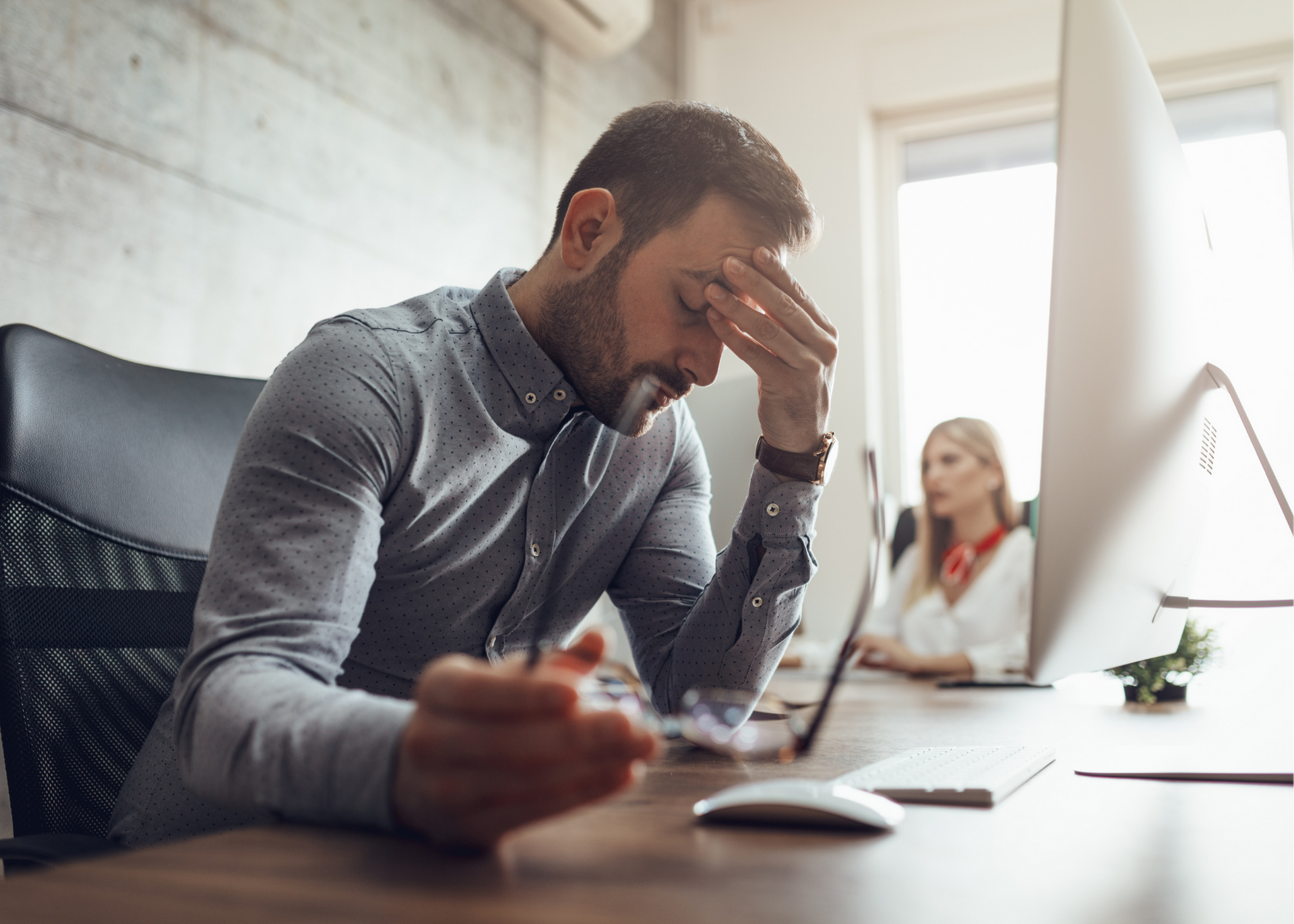 Man holding his head at a desk in a bright office, looking stressed while a blurred coworker sits behind him.