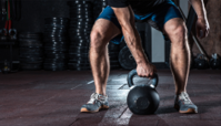 Athlete gripping a kettlebell in a gym, with muscular legs and dumbbells in the background