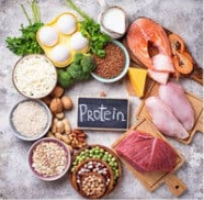 Assorted protein foods on a table, including eggs, fish, chicken, beef, legumes, and grains.