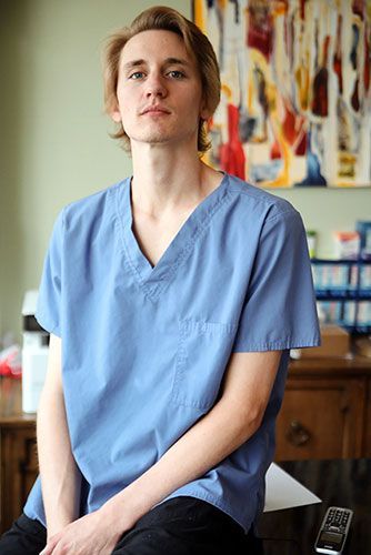 Person in blue scrubs seated indoors, with abstract artwork and shelves in the background.