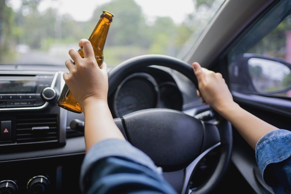 Person driving while holding a beer bottle, seen from inside a car.