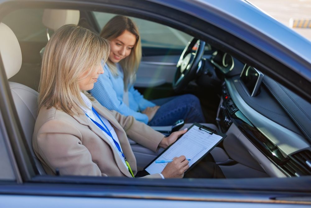 Two people inside a car reviewing paperwork, one writing on a clipboard and the other looking on.
