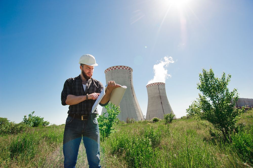 Worker in hard hat holding clipboard near cooling towers under a bright blue sky.