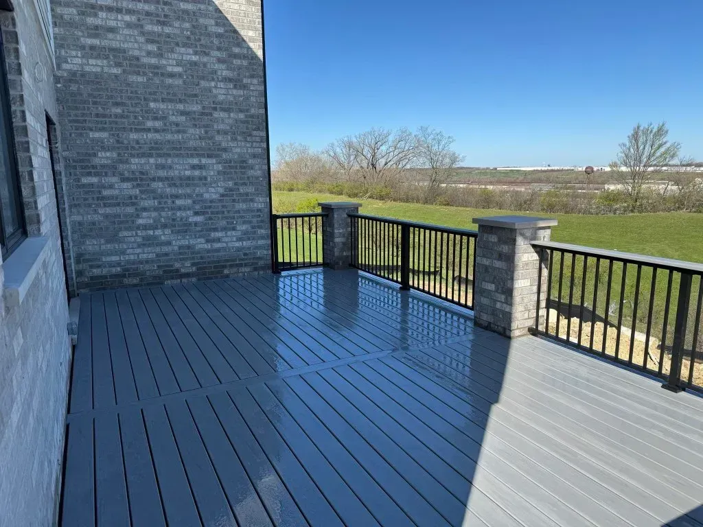 Brick patio deck with black railings beside a house, overlooking a grassy field and blue sky.