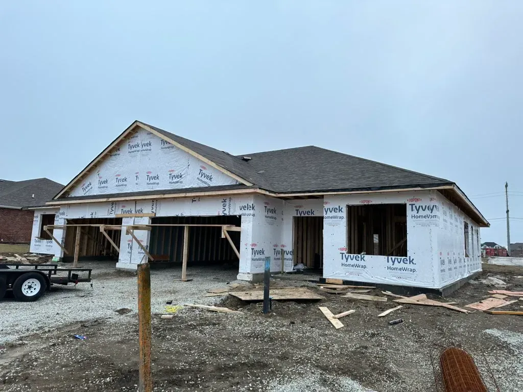 Newly built single-story house under construction with white wrap, gray roof, and muddy lot