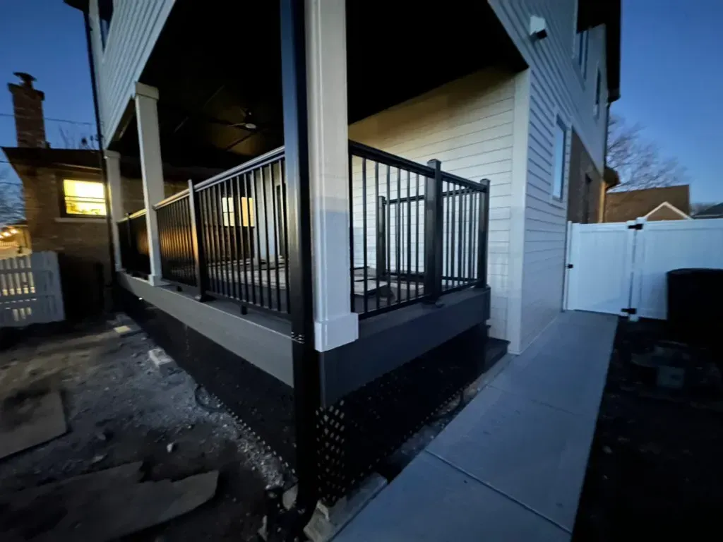 Exterior side porch with black railing and white siding beside a concrete walkway at dusk