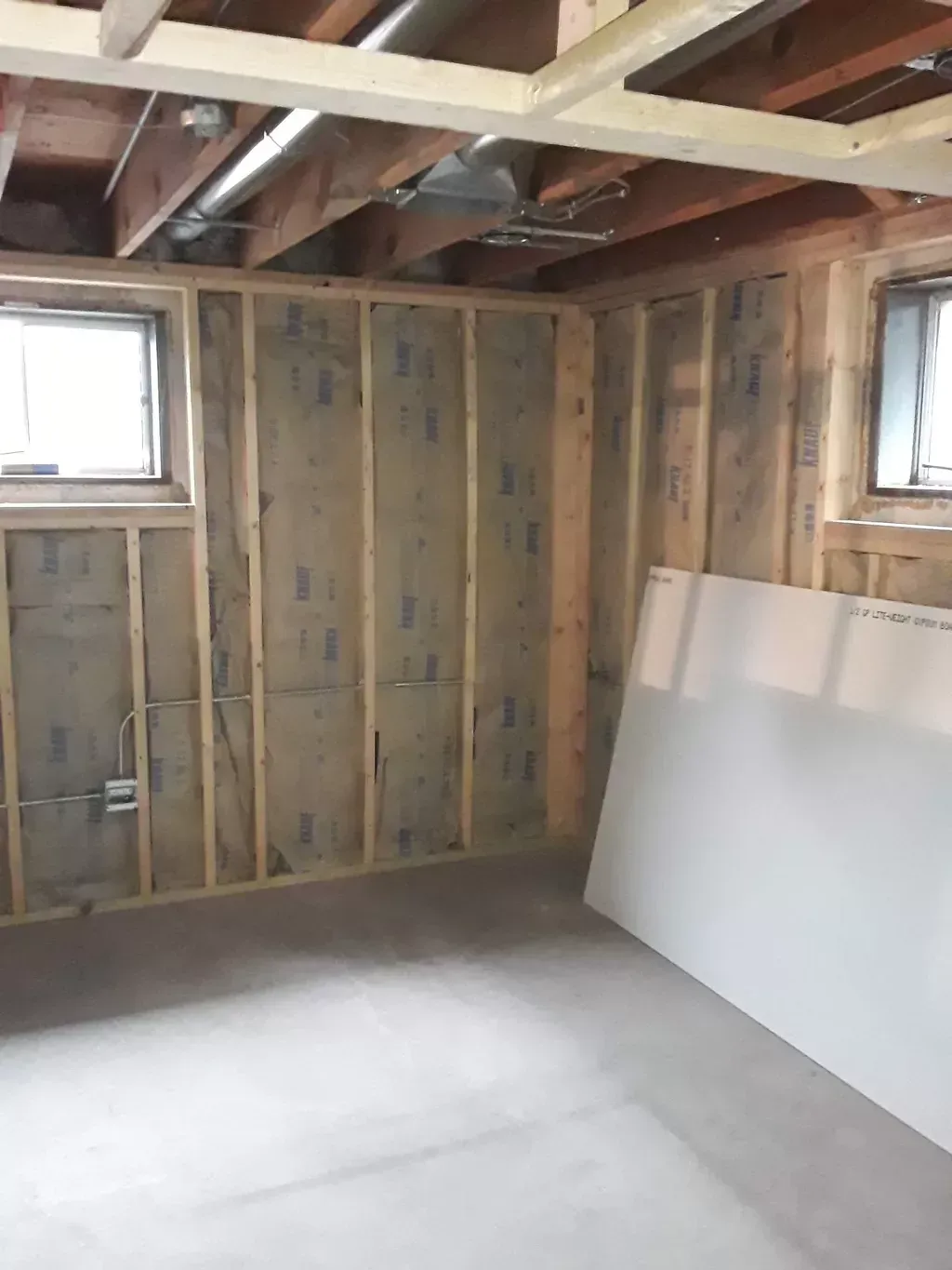 Unfinished basement room with exposed wall studs, ceiling beams, small windows, and drywall leaning against the wall