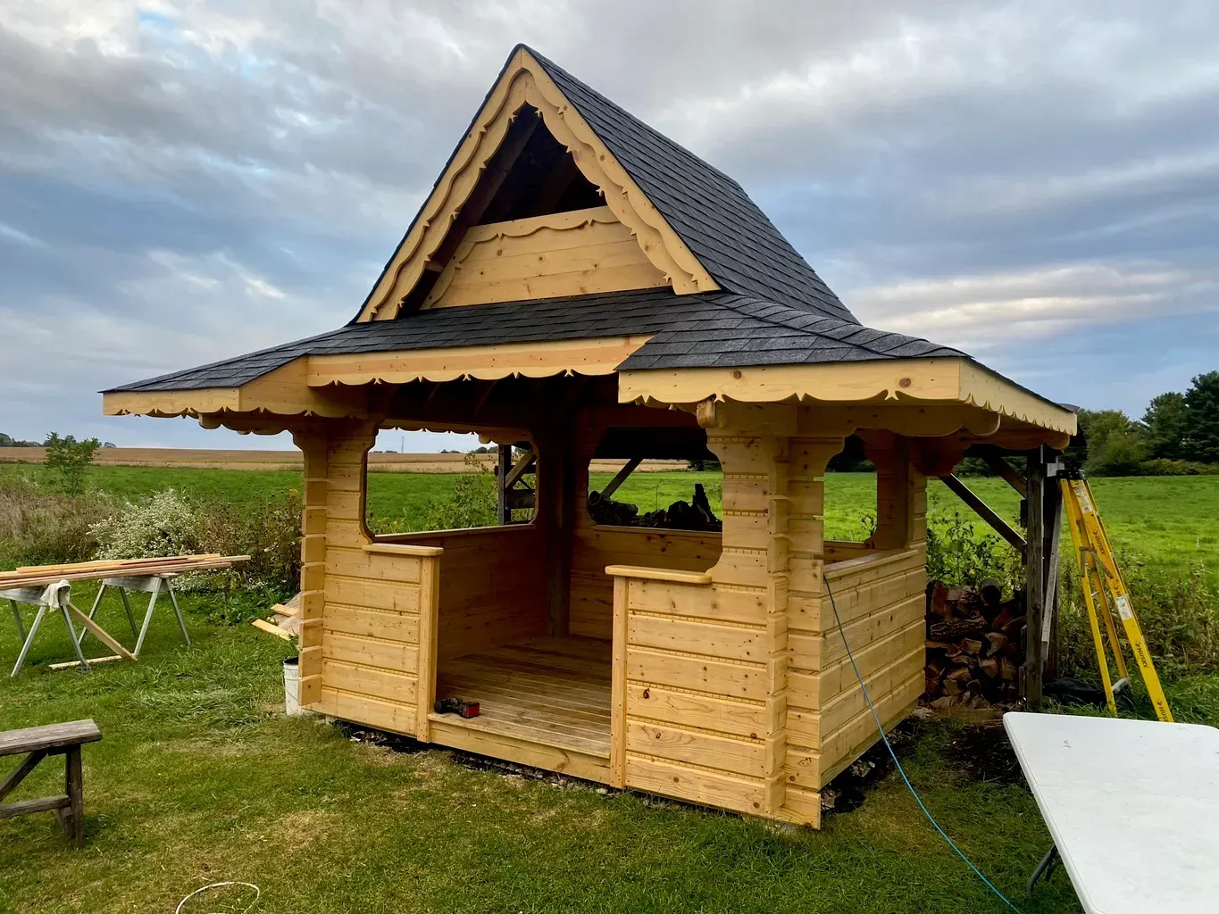 Wooden garden playhouse with dark shingled roof in a grassy field under a cloudy sky