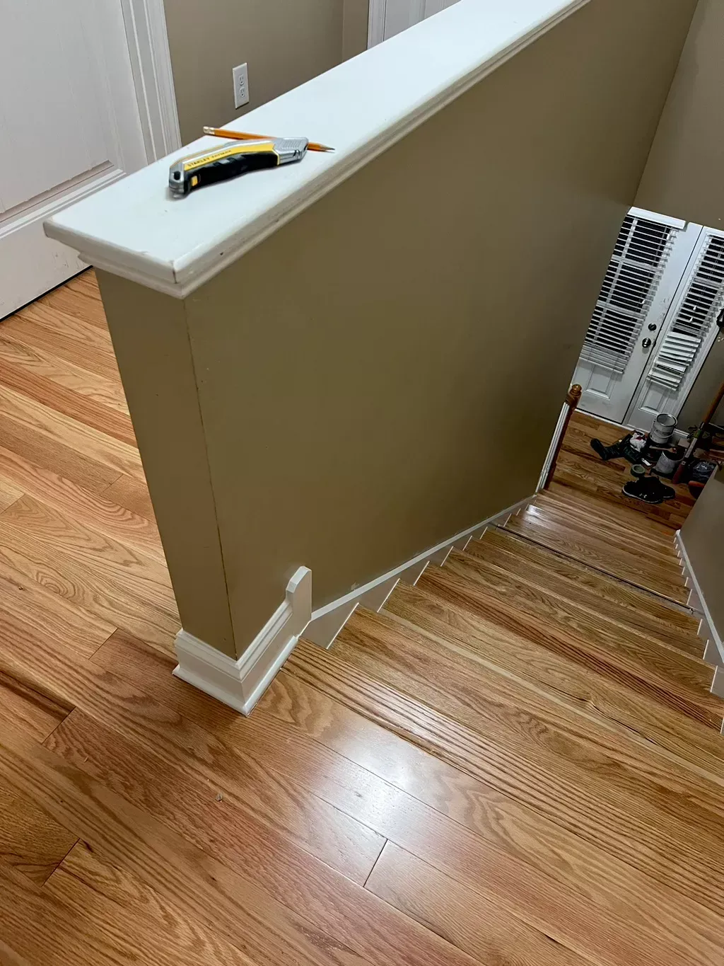 Stair landing with beige half wall, hardwood floor, and a yellow utility knife on the railing.