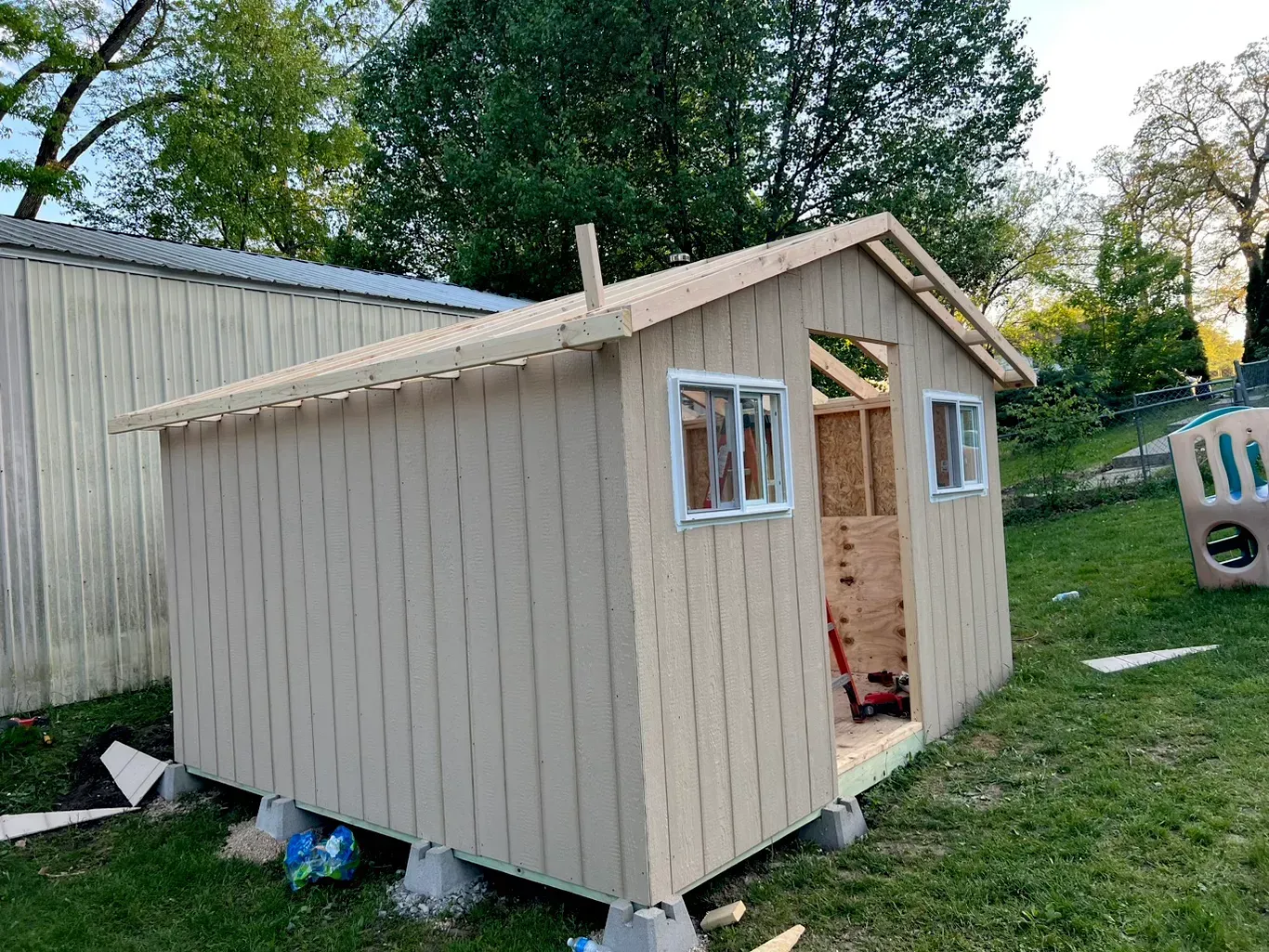 Small unfinished beige shed with white trim in a grassy yard, viewed from the front-left side.