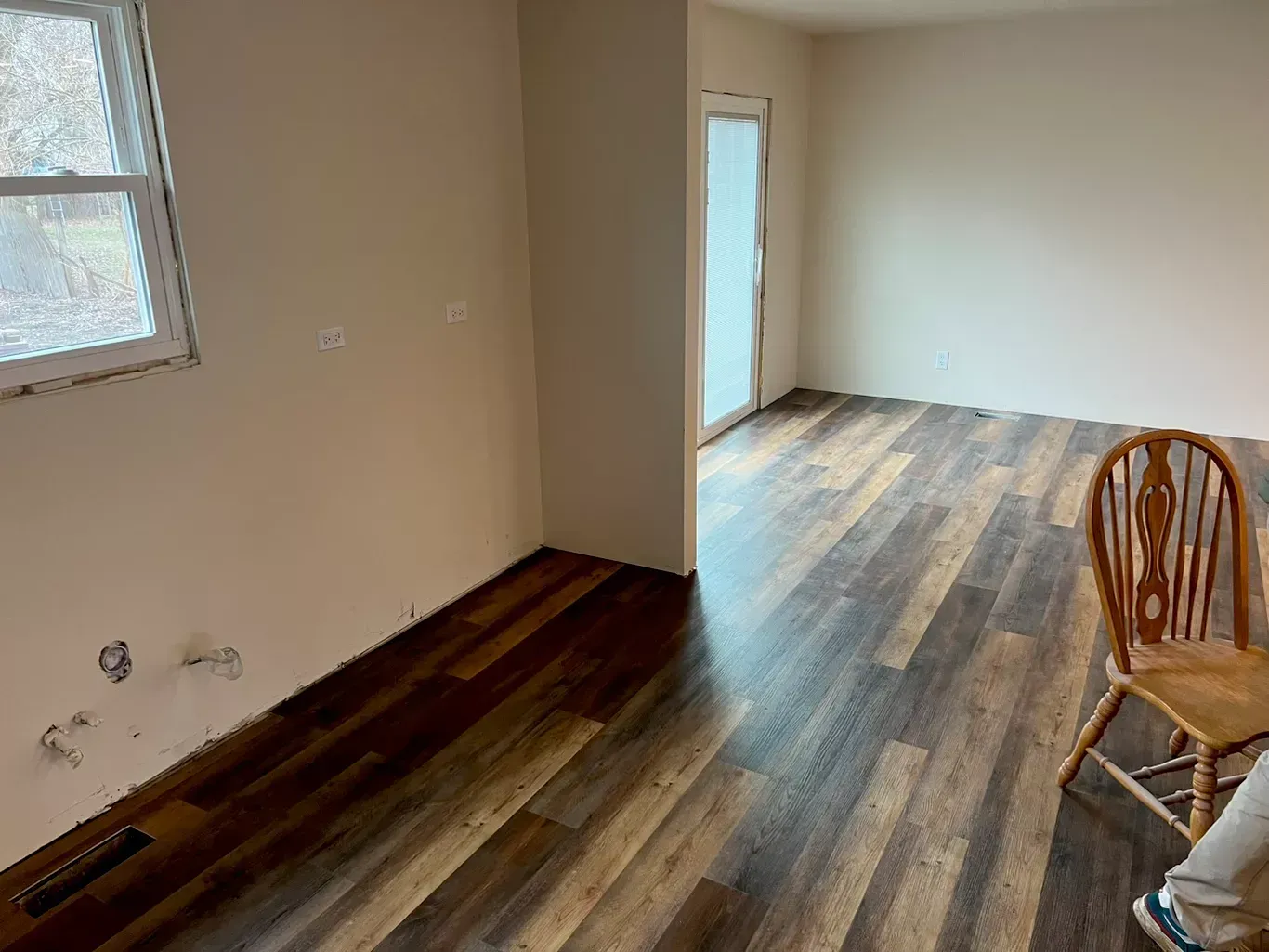Empty room with unfinished wood floors, beige walls, windows, and a wooden chair by the wall