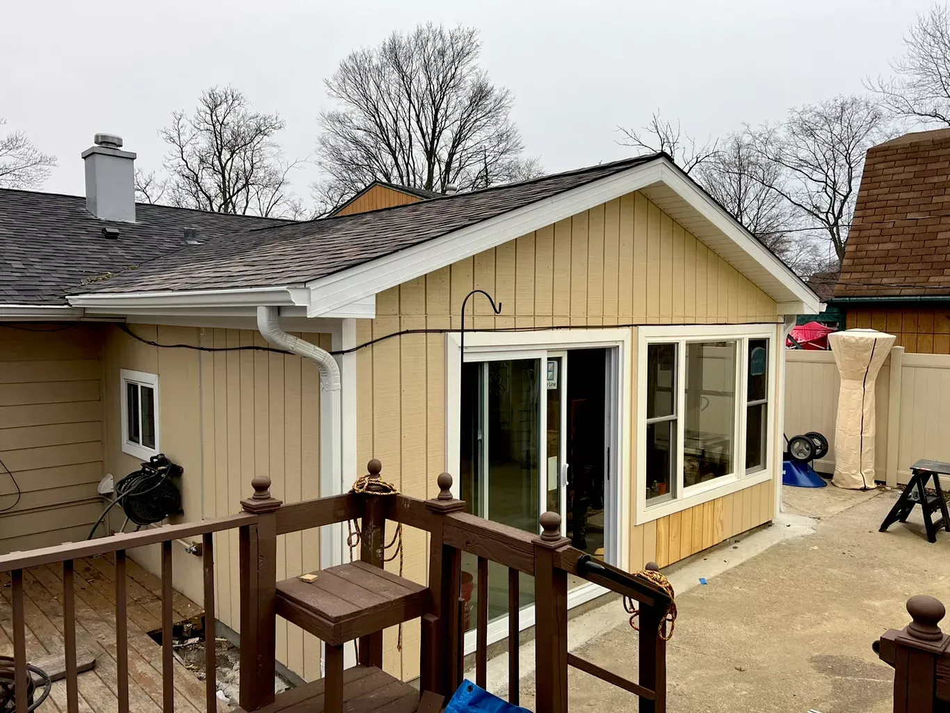 Backyard view of a tan house with white trim, deck railing, and a small patio under cloudy skies