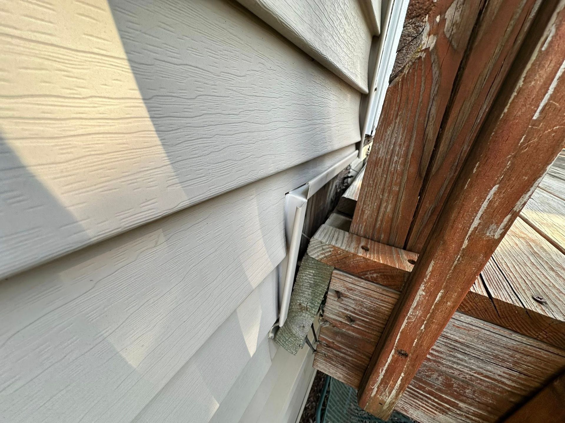 Close-up of weathered wooden trim beside white house siding, showing a damaged gap and exposed flashing