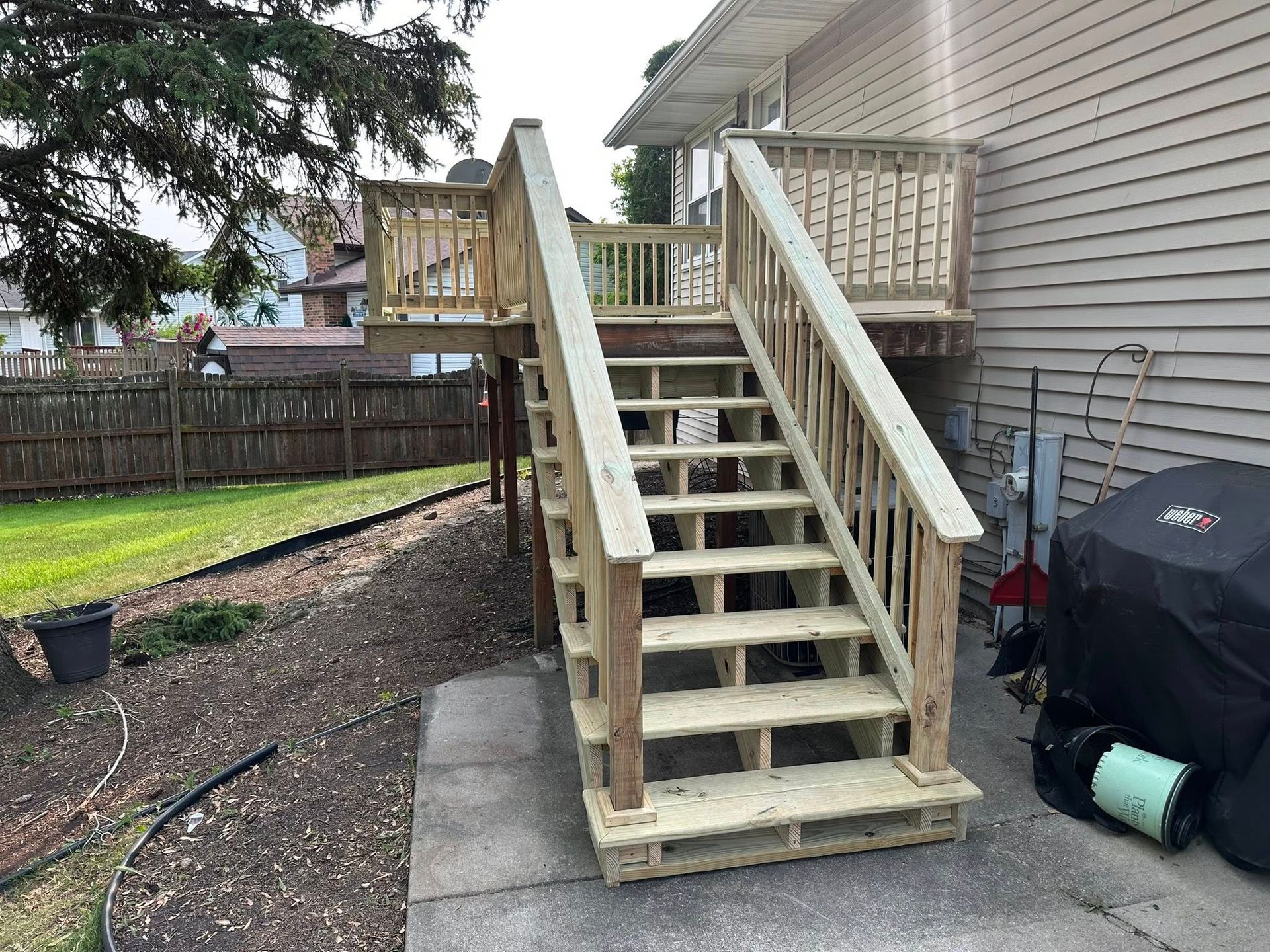 Wooden outdoor staircase leading to a raised deck beside a house, with a grill and fenced yard.
