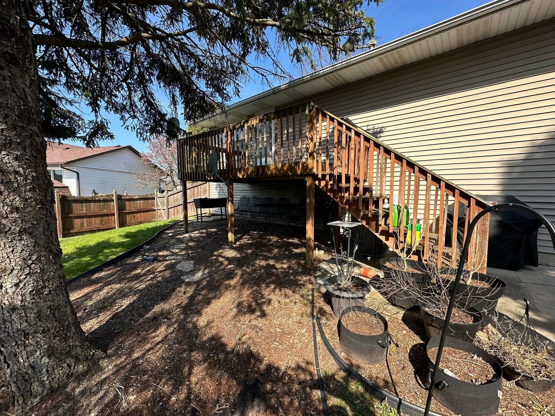 Backyard deck with wooden stairs, beige house siding, patio chairs, and a large tree in the foreground