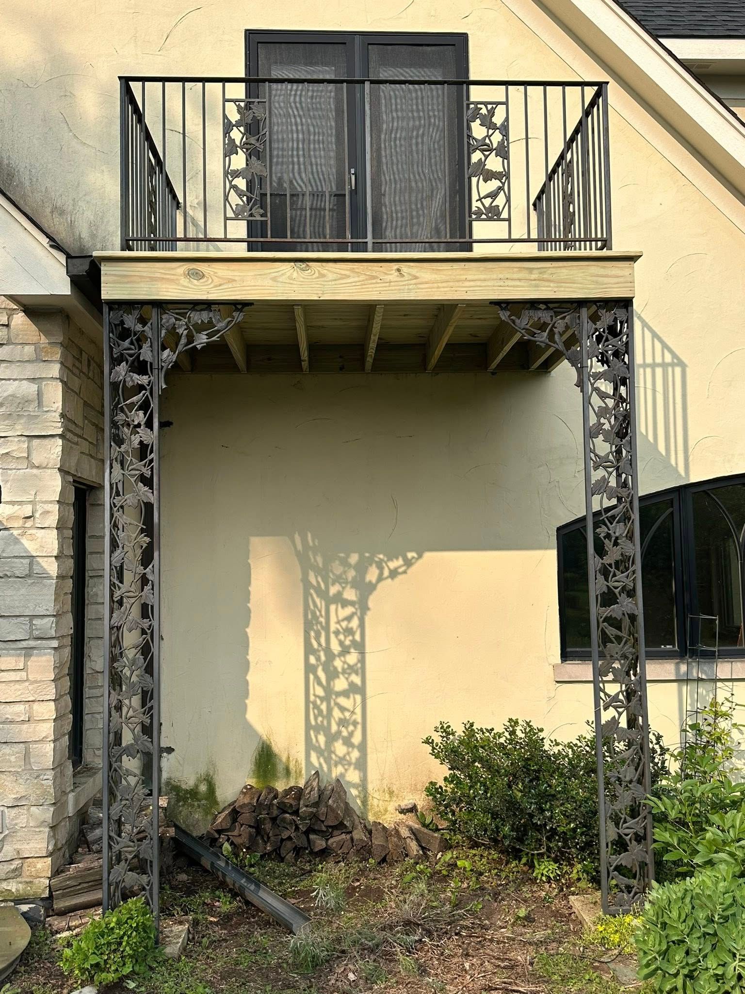 Apartment balcony with black railings, stone columns, and beige walls above a small garden.