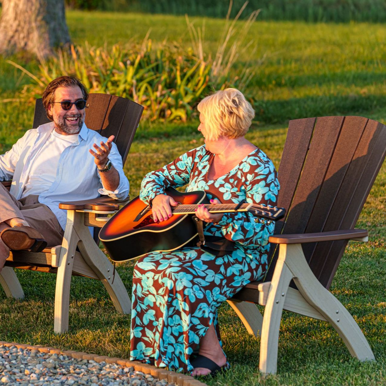Two people sit in outdoor chairs on grass; one plays an acoustic guitar while the other gestures during a conversation.